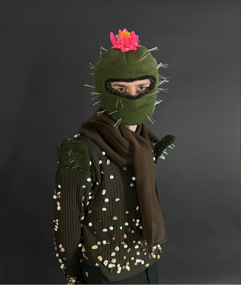 Studio photo of the Superfluous Design look: green balaclava with pins and a pink flower, pistachio shells scattered on dark knit.