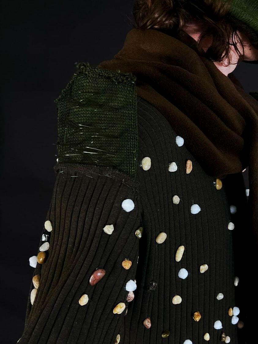 Close-up of the Superfluous Design shoulder: burlap patch, pins, and pistachio shells on dark knit.